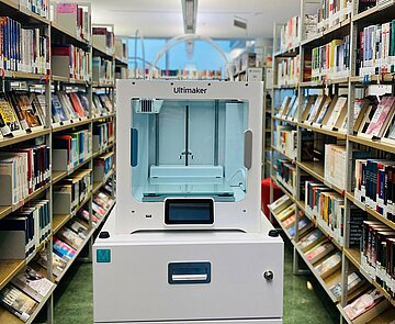 Der 3D-Drucker steht inmitten der Bibliothek zwischen Bücherregalen