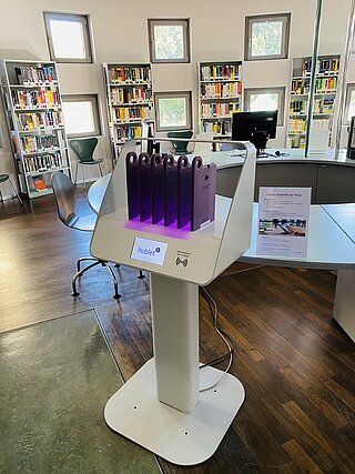 Ein Foto von unserem Hublet-Ständer, in dem sechs Tablets mit lila Hüllen bereits zur Ausleihe stehen. Im Hintergrund ist die Computerbibliothek mit einem Rechner, Sitzplätzen und vollen Bücherregalen zu sehen.