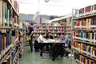 Eine gruppe junger Männer sitzt an einem Gruppentisch im Neuen Ständehaus zwischen Bücherregalen und arbeitet.