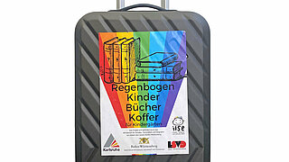 Der Regenbogenkinderbücherkoffer vor weißem Hintergrund. Ein Rollkoffer mit einem Rgeenbogenplakat drauf.